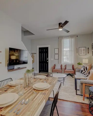Open-plan living and dining area with wood table, gray sofa, wall-mounted TV, and staircase visible in background