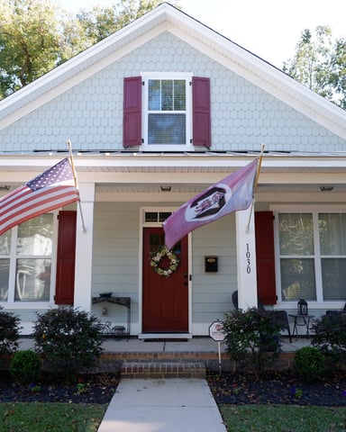 White cottage with red shutters, front porch with columns, American flag and purple flag, wreath on red door, surrounded by shrubs and trees