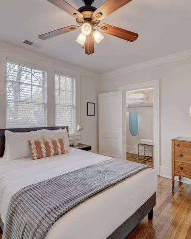 Bright bedroom with queen bed, wood dresser, shuttered windows, ceiling fan with lights, and hardwood floors.