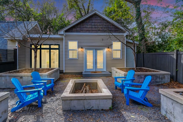 Modern cottage with gray siding and blue doors, surrounded by blue Adirondack chairs and concrete fire pit in gravel courtyard at dusk.