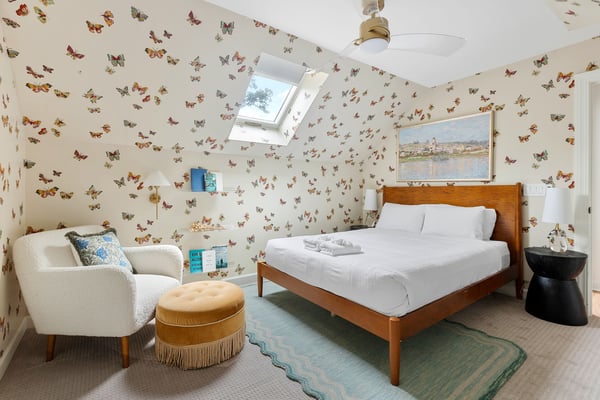 Attic bedroom with butterfly-patterned wallpaper, wood bed with white linens, cream armchair, and skylight window.