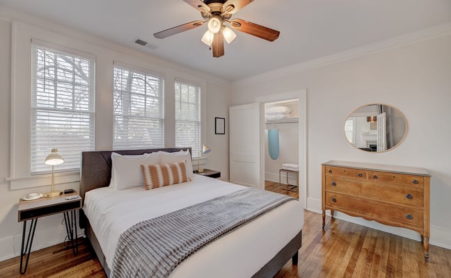 Furnished bedroom with warm hardwood floors, white bedding, antique dresser, and ceiling fan in a Heartwood monthly rental