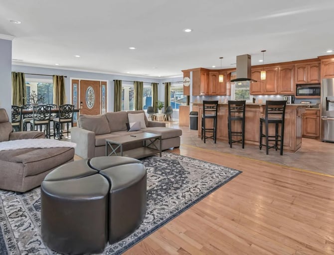 Open-plan living room and kitchen with wood flooring, gray seating, wood cabinetry, and water views through large windows